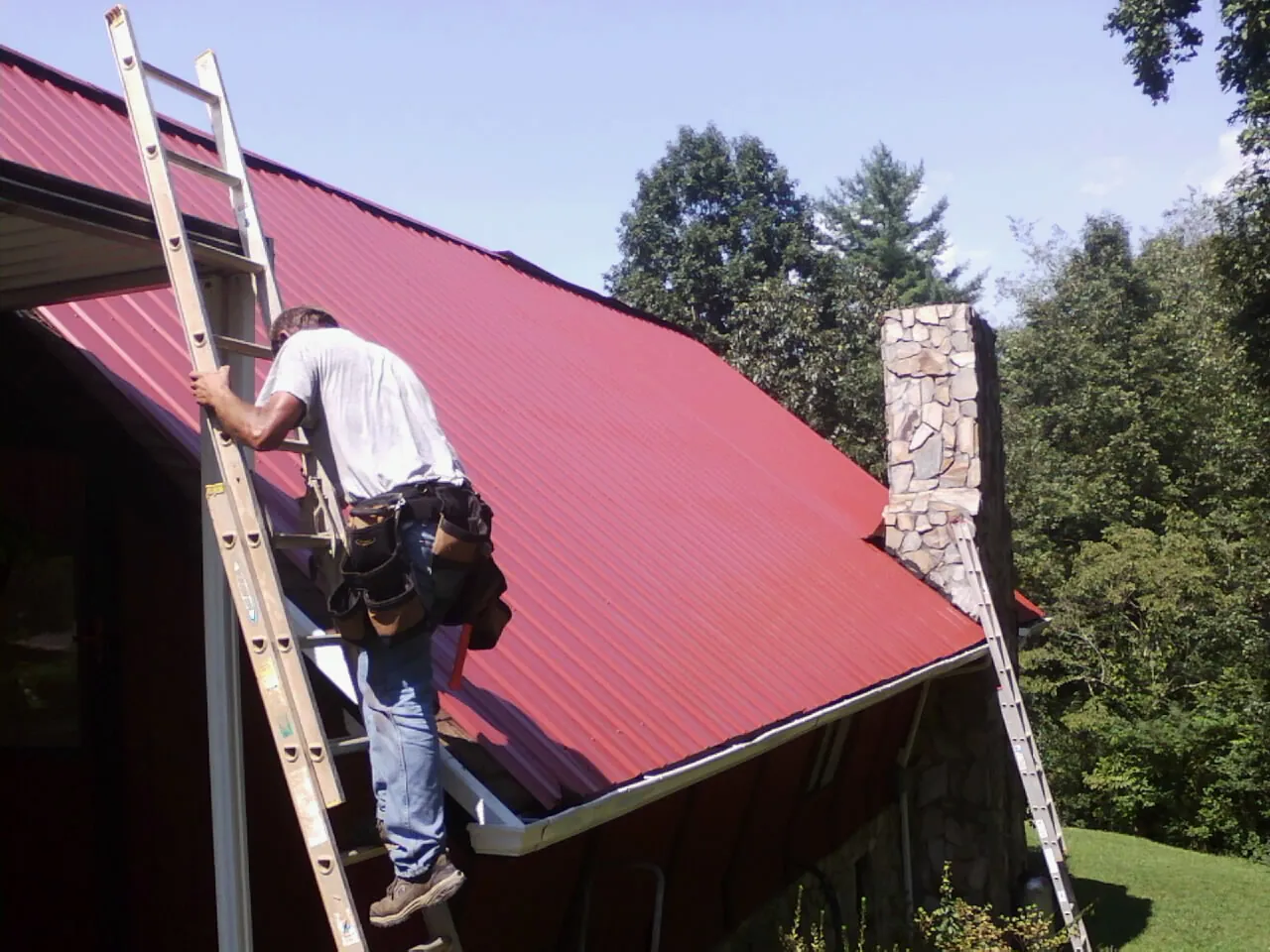 Roofer on a ladder beside a red metal roof and stone chimney — Soffit & Fascia Repair by Roofing Brothers in Fort Deposit